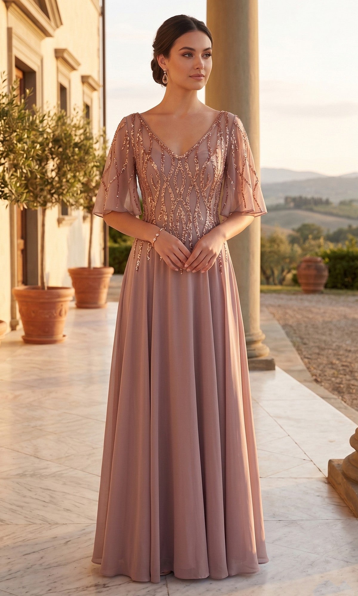 A woman in the Chicas J064 Long Prom Dress, a mocha and gold floor-length chiffon A-line gown with beaded details, stands on a marble patio near pillars and potted plants at sunset.