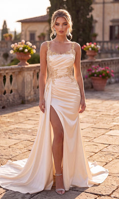 A woman wearing the elegant Chicas J092 long prom dress in champagne, featuring gold floral embroidery and a high leg slit, stands outdoors on a stone terrace adorned with potted flowers and historic buildings in the background.