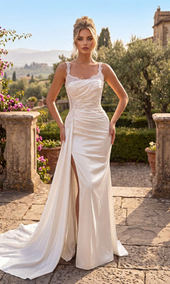 An elegant woman stands outdoors on a stone terrace surrounded by greenery and flowers, wearing the Chicas J092 Long Prom Dress in off white—a satin gown with lace straps, beautiful floral embroidery, and a stylish thigh-high leg slit.