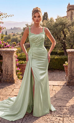 A woman stands on a stone terrace surrounded by greenery and distant hills, wearing the Chicas J092 Long Prom Dress in sage—a light green satin gown with lace details, subtle floral embroidery, and a thigh-high slit that highlights her elegance.