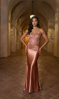 A woman stands on a tiled floor beneath arched ceilings with soft lighting, wearing the Chicas J094 long prom dress in rose gold—a shiny, off-the-shoulder gown featuring a satin sheath skirt and an elegant lace corset bodice.