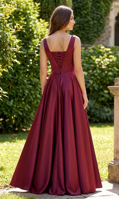 A woman stands outdoors in a garden, wearing the Chicas J112 long prom dress—a sleeveless, floor-length burgundy satin gown featuring a lace-up back.