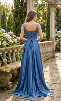 A woman wearing the Long Prom Dress: Chicas J112, a blue satin gown with a lace-up back, stands outdoors on a stone path near flowers and columns, facing away from the camera.