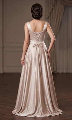 A woman wearing the Long Prom Dress: Chicas J112, an elegant floor-length satin gown with a lace-up back, stands on a wooden floor in a formal, neutral-toned room.