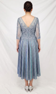 A woman stands facing away, wearing the Modest Midi Wedding Guest Dress: Chicas J152—a silver-blue, knee-length A-line midi dress with a V-back and sheer sleeves—paired with silver shoes against a plain white background, creating a chic and elegant look for any wedding guest.