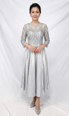 A woman stands against a white backdrop, wearing the Modest Midi Wedding Guest Dress: Chicas J152 in silver—an elegant a-line midi dress featuring lace detailing and three-quarter sleeves—paired with matching silver heels, making it the perfect choice for a sophisticated wedding guest ensemble.