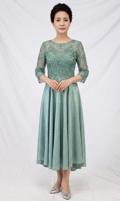 A woman stands in front of a white backdrop wearing the Modest Midi Wedding Guest Dress: Chicas J152 in sage, featuring lace and satin fabric, an A-line midi silhouette with three-quarter sleeves, paired with silver shoes.