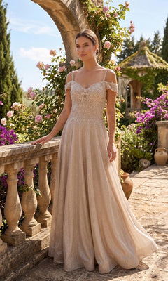 A woman in a champagne Long Prom Dress: Chicas J169 with an off-shoulder lace design and embellished bodice stands on a stone terrace surrounded by blooming flowers and greenery on a sunny day.