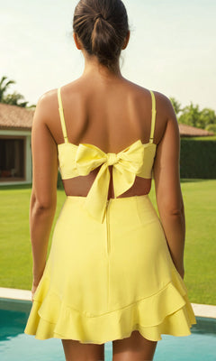 A woman stands by a pool, facing away from the camera, wearing the affordable Tie-Back Short Yellow Party Dress J344894, with a large bow at the back. A lawn and house are visible in the background.