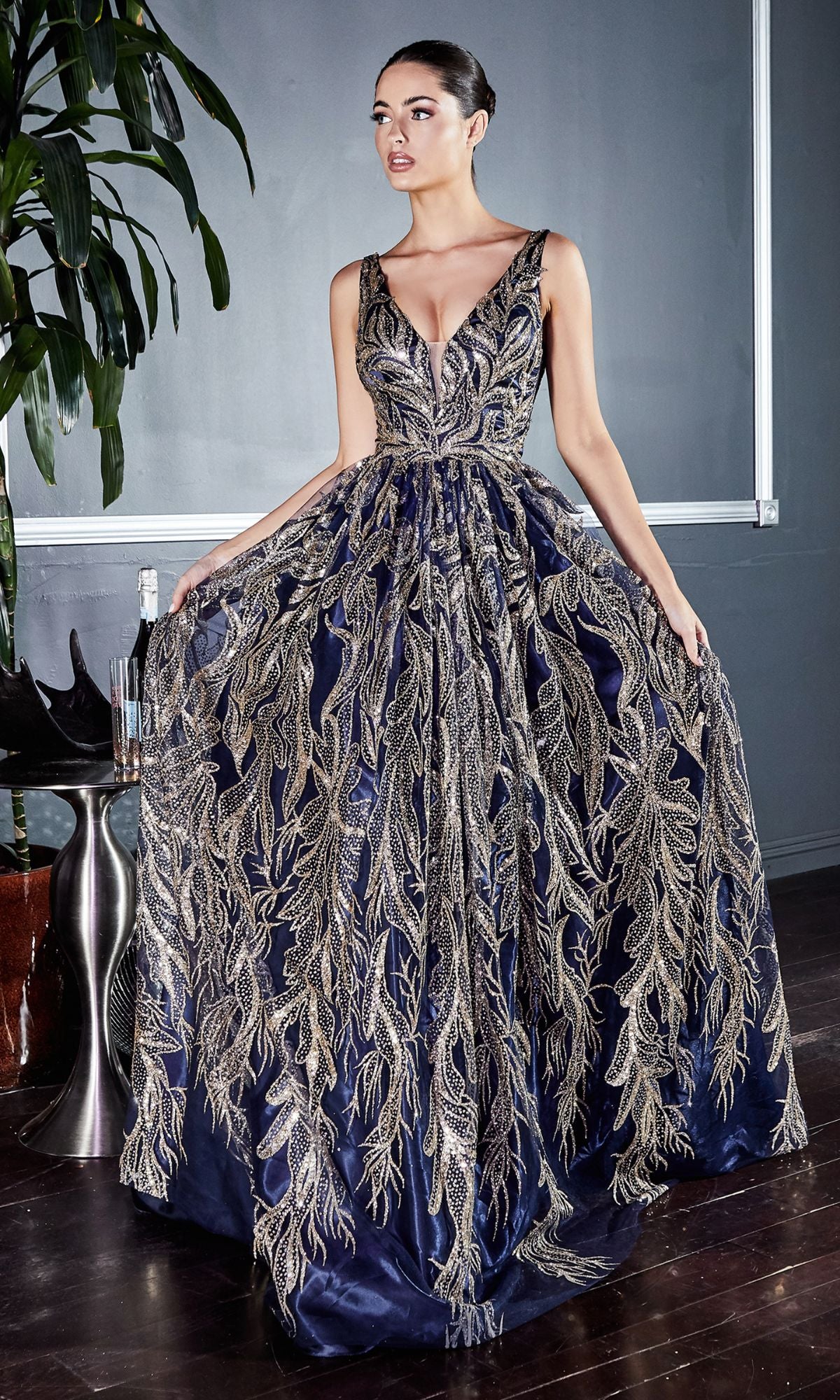 A woman stands indoors in the Sleeveless Glitter-Print Long Prom Dress J812, holding the sides of the skirt to showcase its intricate navy blue and gold glitter detailing.