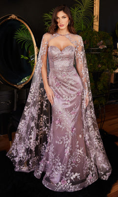 A woman stands indoors wearing the Strapless Long Pageant Dress with Cape J834, a fitted lavender gown featuring a sweetheart neckline and sheer, embroidered floral cape, in front of a mirror and greenery.