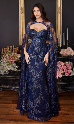 A woman stands in front of a black dresser with flowers and candles, wearing the Strapless Long Pageant Dress with Cape J834—navy blue, strapless, long, with floral embroidery, a sweetheart neckline, and a matching sheer cape.