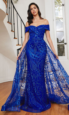 Indoors, a woman wears the Long Prom Dress with Glitter-Print Overskirt J836, an off-the-shoulder royal blue gown featuring sheer overlay panels adorned with baroque glitter prints.