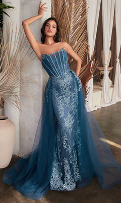 A woman poses indoors in the Glitter-Print Strapless Long Prom Dress J858 in lapis blue, featuring lace details and a sheer overlay, surrounded by neutral-toned dried palm leaves.