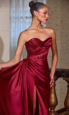 A woman wearing the Ladivine J887 Side-Drape Strapless Long Prom Gown in burgundy satin with a sequined bodice and high slit stands by a staircase, holding part of her elegant evening gown.