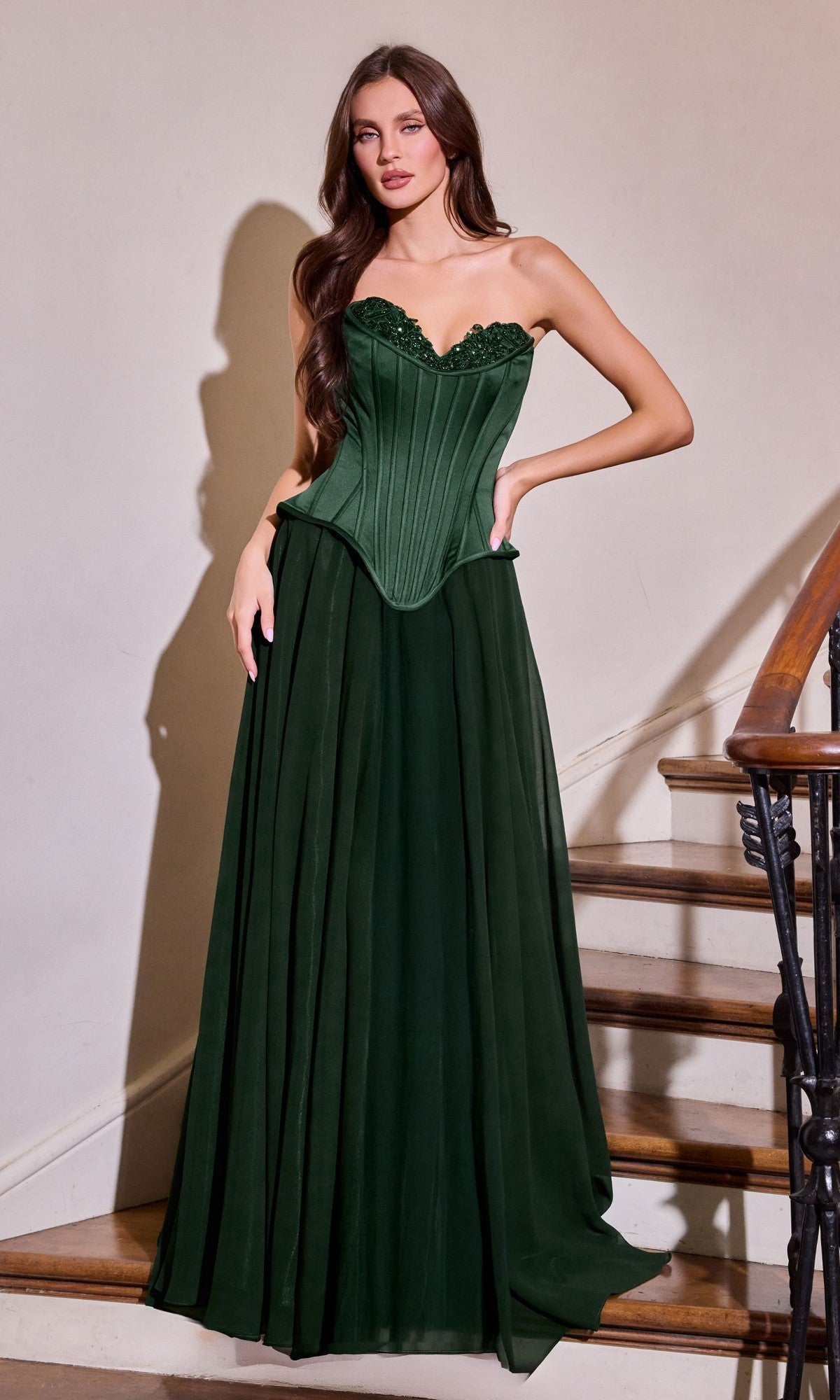A woman stands indoors near a staircase, wearing the Ladivine J891 long prom dress in emerald, featuring a strapless corset bodice and a flowing chiffon A-line skirt.