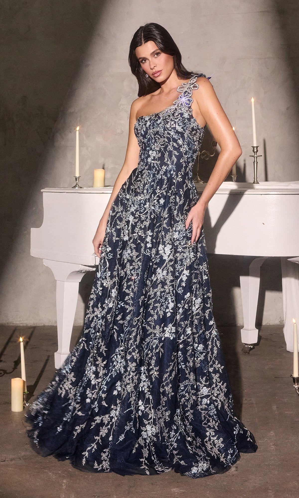 A woman in the navy Ladivine J909 long prom dress poses by a white piano, candlelight illuminating her elegant one-shoulder floral embroidery and silver accents.