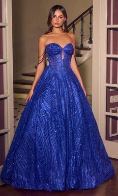 A woman in the Ladivine J915 long prom dress in royal stands indoors near a staircase, wearing a strapless shimmering blue gown with delicate crystal beadwork; her hair is down and her arms rest at her sides.