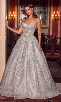 A woman stands indoors in an elegant, dimly lit room wearing the Ladivine J915 long prom dress in silver—a strapless ball gown featuring a fitted bodice embellished with delicate crystal beadwork and a full, shimmering skirt of glitter tulle.