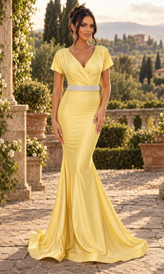 A woman stands outdoors on stone pavement, surrounded by greenery and distant hills, wearing the Jessica Angel JA879 long prom dress in pale yellow. The floor-length gown features short sleeves and a silver waistband, styled elegantly for a mother-of-the-bride look.