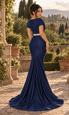 A woman stands outdoors on a stone terrace, facing away, wearing the fitted, floor-length Jessica Angel JA879 navy blue long prom dress with a train; greenery and distant buildings appear in the background.