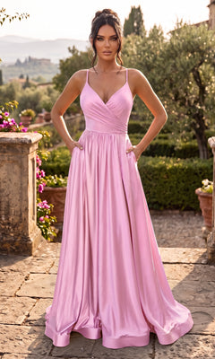 A woman in the Jessica Angel JA894 Long Prom Dress in Bubblegum, featuring a lace-up strappy back, stands outdoors on a stone patio surrounded by greenery and flowers, with distant hills in the background.