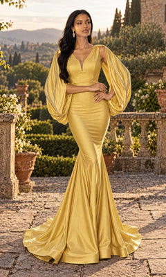 A woman stands outdoors on a stone terrace with greenery and mountains in the background, wearing the Jessica Angel JA902 long prom dress in gold, featuring a fitted mermaid silhouette and dramatic draped sleeves.