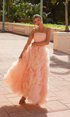 A woman stands outdoors on a sunlit walkway, greenery behind her, wearing the Nox Anabel JQ1037 Strapless Peach Prom Ball Gown with a voluminous ruffled skirt.