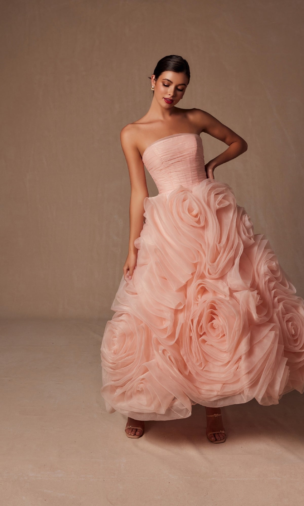 A woman models the Nox Anabel JQ1037, a strapless peach prom ball gown featuring oversized rosettes on the full skirt, set against a neutral background.