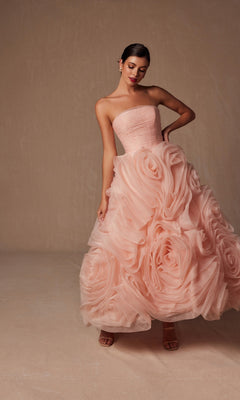 A woman models the Nox Anabel JQ1037, a strapless peach prom ball gown featuring oversized rosettes on the full skirt, set against a neutral background.