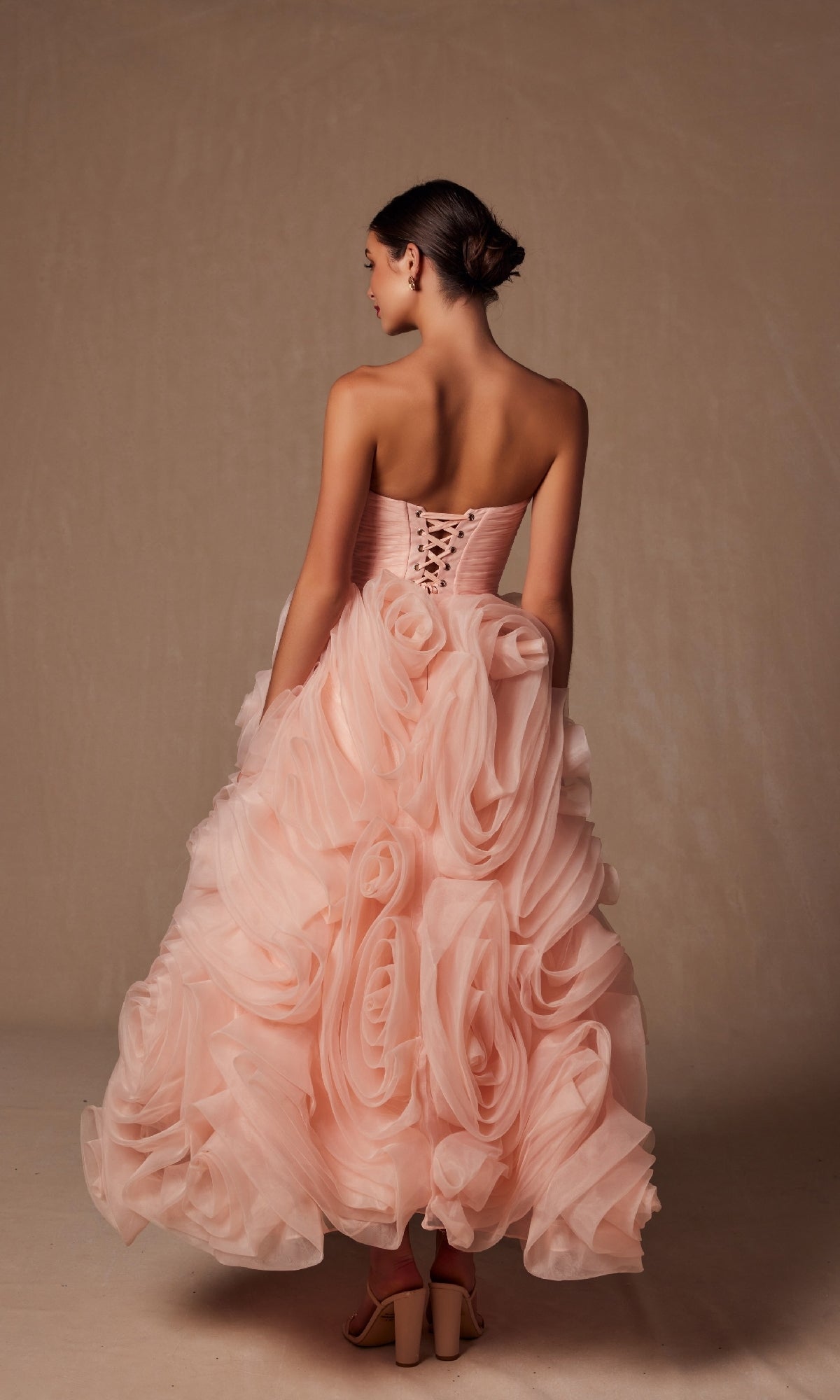 A woman stands facing away, wearing the Nox Anabel JQ1037 strapless peach prom ball gown with a corset back and a cascading ruffled skirt featuring oversized rosettes.