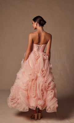 A woman stands facing away, wearing the Nox Anabel JQ1037 strapless peach prom ball gown with a corset back and a cascading ruffled skirt featuring oversized rosettes.