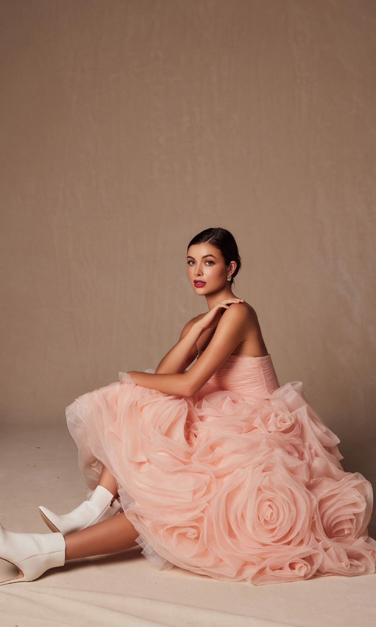 A woman sits on the floor in the Nox Anabel JQ1037 strapless peach prom ball gown with oversized rosettes and white ankle boots, against a plain beige background.