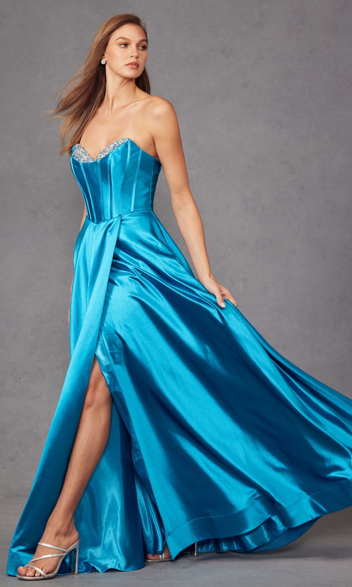 A woman poses against a plain gray background in the Strapless Long Blue Prom Dress: Juliet JT2435A, featuring a shiny blue fabric, sweetheart neckline, corset-style bodice, and high slit.