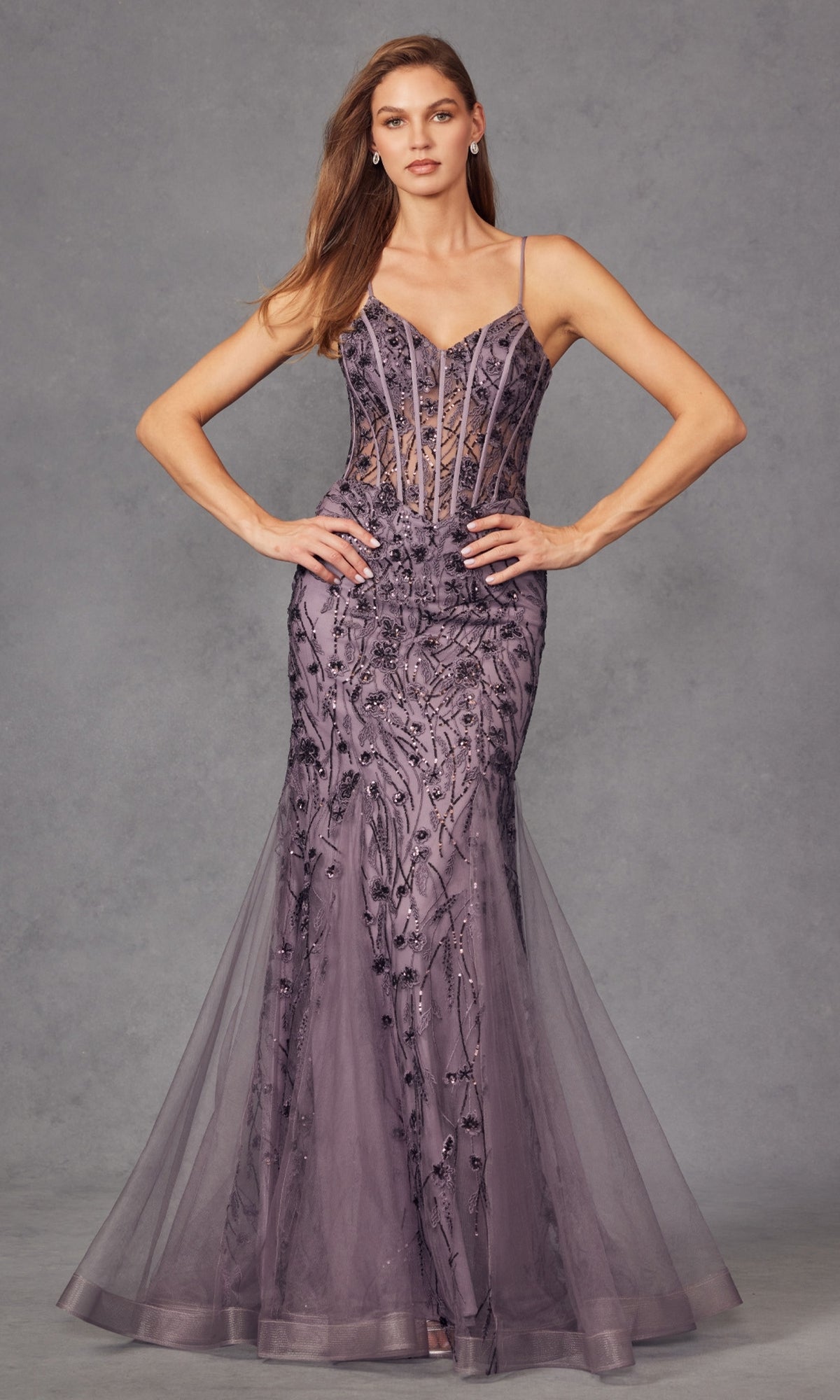 A woman stands with hands on hips wearing the Long Sequin Mermaid Prom Dress: Juliet JT2441S, a floor-length gown with sheer bodice, thin straps, and intricate sequin details against a gray studio background.
