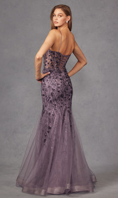 A woman poses in the Juliet JT2441S Long Sequin Mermaid Prom Dress, featuring a floor-length lavender design with thin straps, sheer panels, and intricate embroidery, set against a gray studio backdrop.