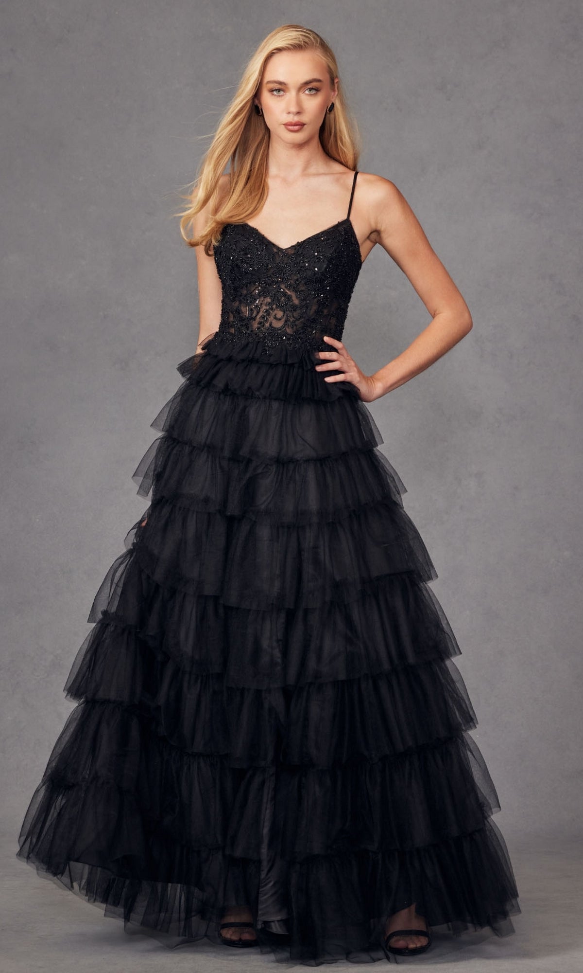 A woman wearing the Juliet JT2451S Long Ruffled Prom Ball Gown, featuring spaghetti straps and a fitted, semi-sheer embellished bodice, poses against a gray background.