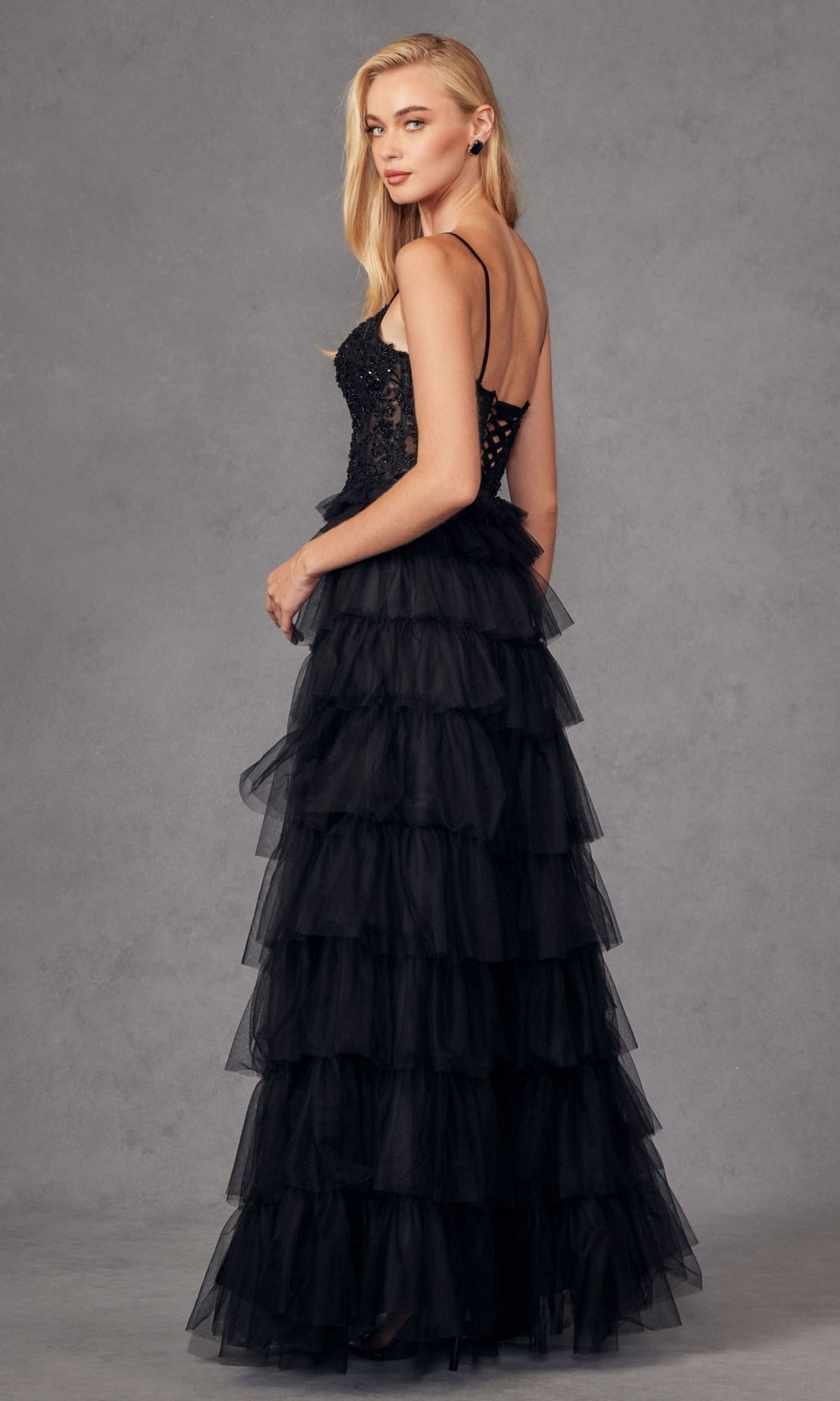 A woman poses against a gray studio background in the Juliet JT2451S Long Ruffled Prom Ball Gown, featuring a black sleeveless design with a tiered tulle skirt and lace bodice.
