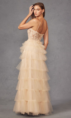 A woman in the Juliet JT2451S sleeveless, floor-length beige ruffled prom ball gown with a lace bodice and layered tulle skirt stands against a gray background, facing away but looking over her shoulder.