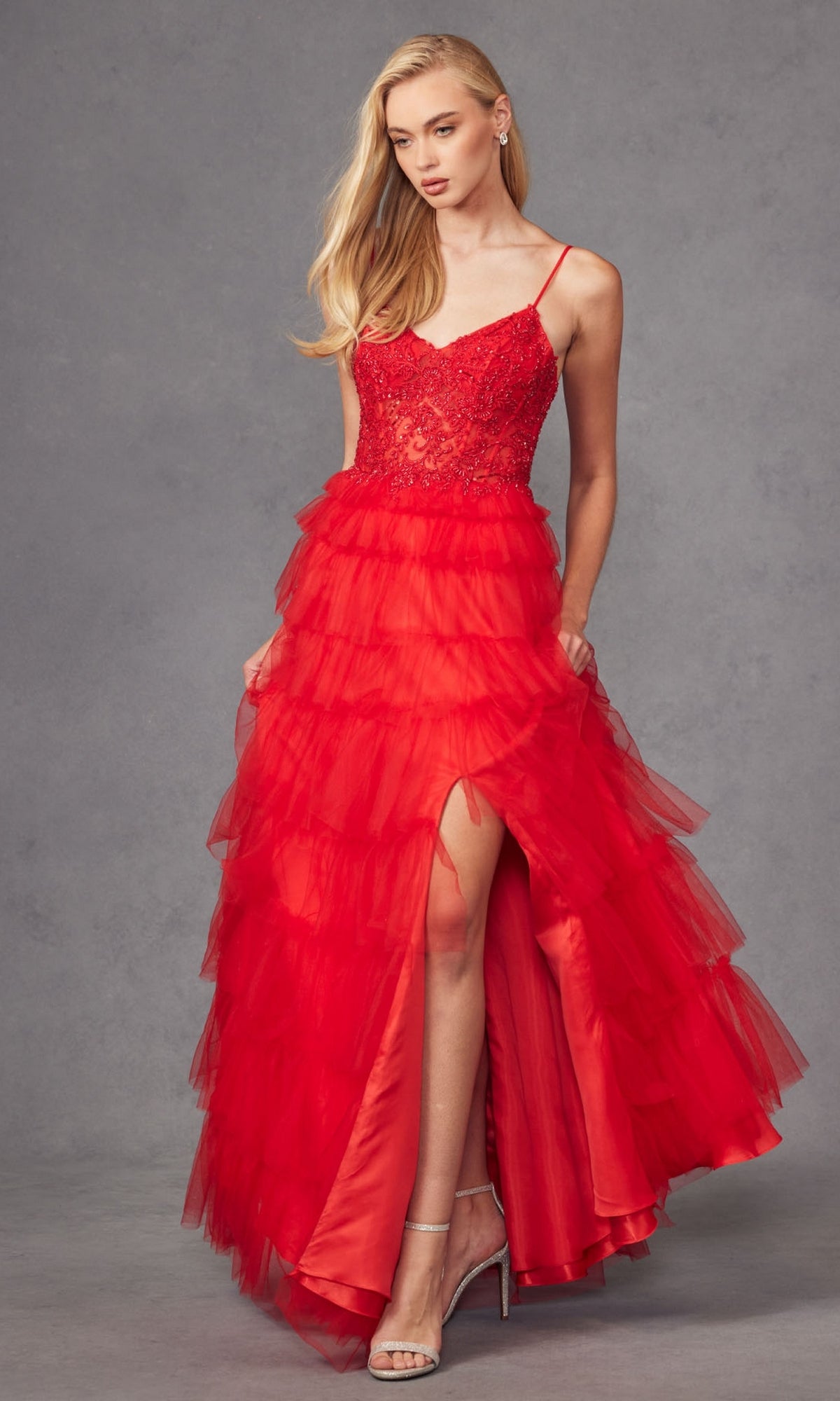 A woman models the Juliet JT2451S Long Ruffled Prom Ball Gown in red tulle with spaghetti straps, a lace bodice, and high slit, posing against a plain grey background.