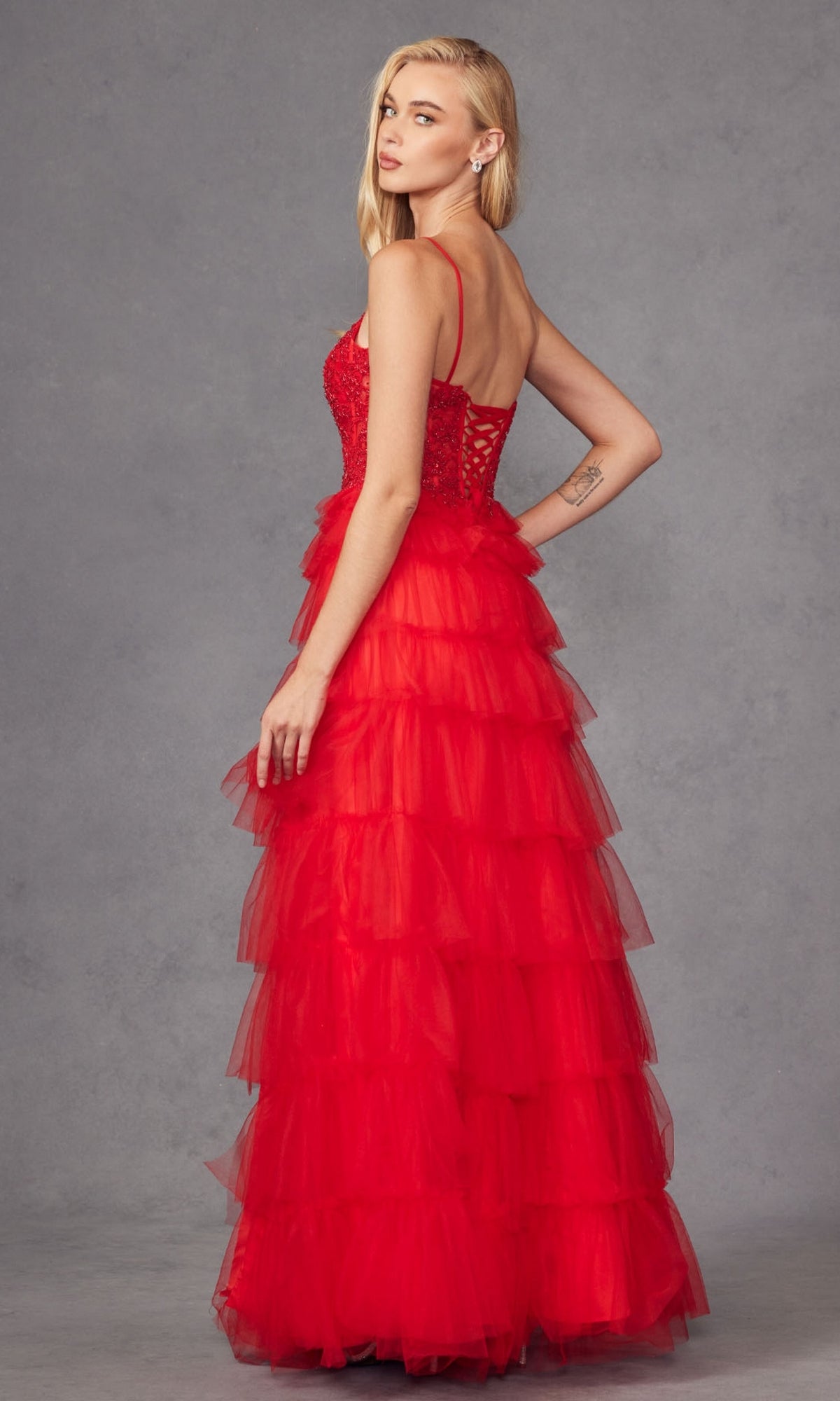 A woman wears the Juliet JT2451S Long Ruffled Prom Ball Gown, featuring a sleeveless red tulle design with a tiered, ruffled skirt, as she poses against a gray background, looking over her shoulder.