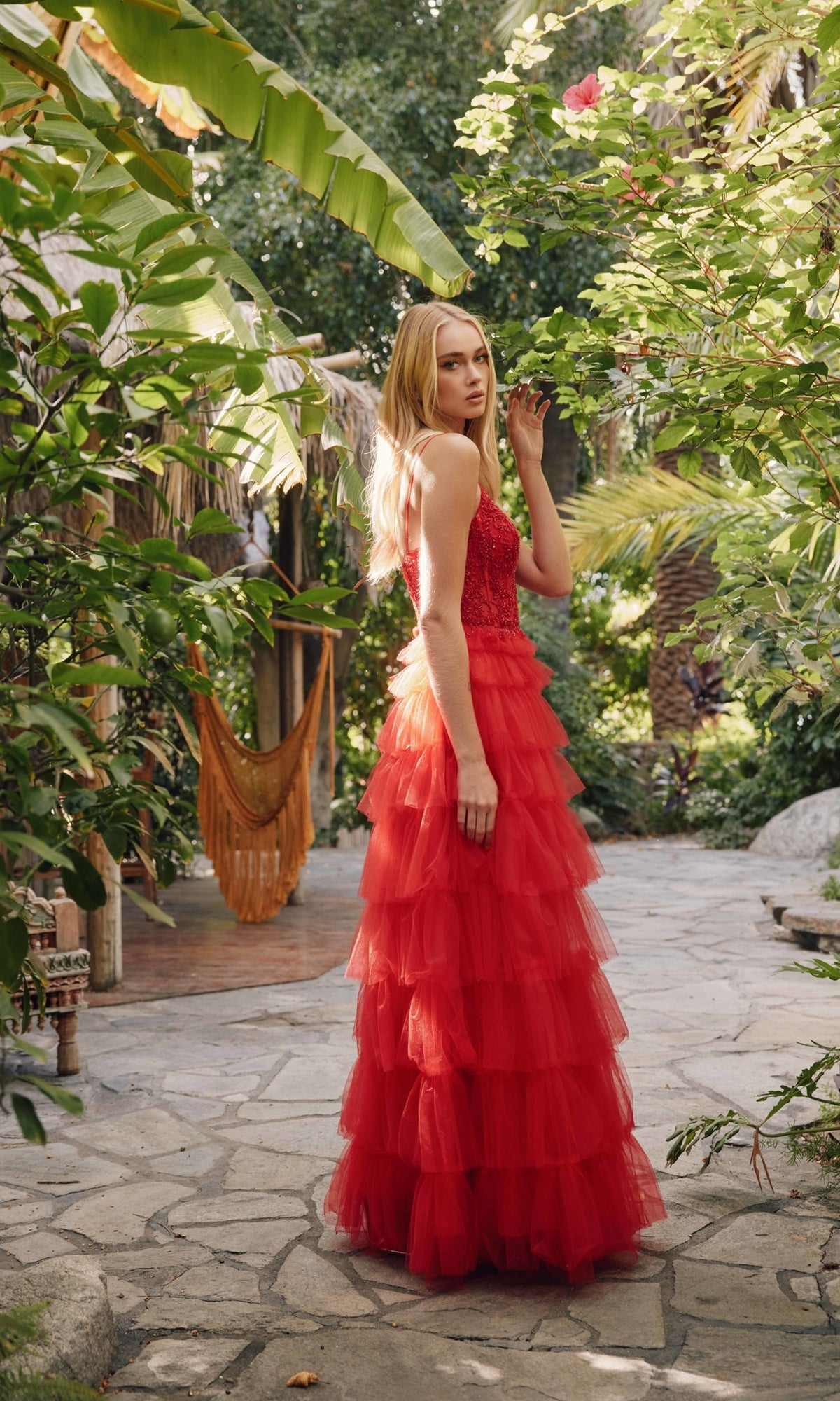 A woman wearing the Long Ruffled Prom Ball Gown: Juliet JT2451S stands on a stone path amid lush green foliage and tropical plants.