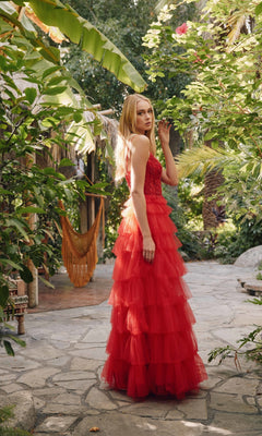 A woman wearing the Long Ruffled Prom Ball Gown: Juliet JT2451S stands on a stone path amid lush green foliage and tropical plants.