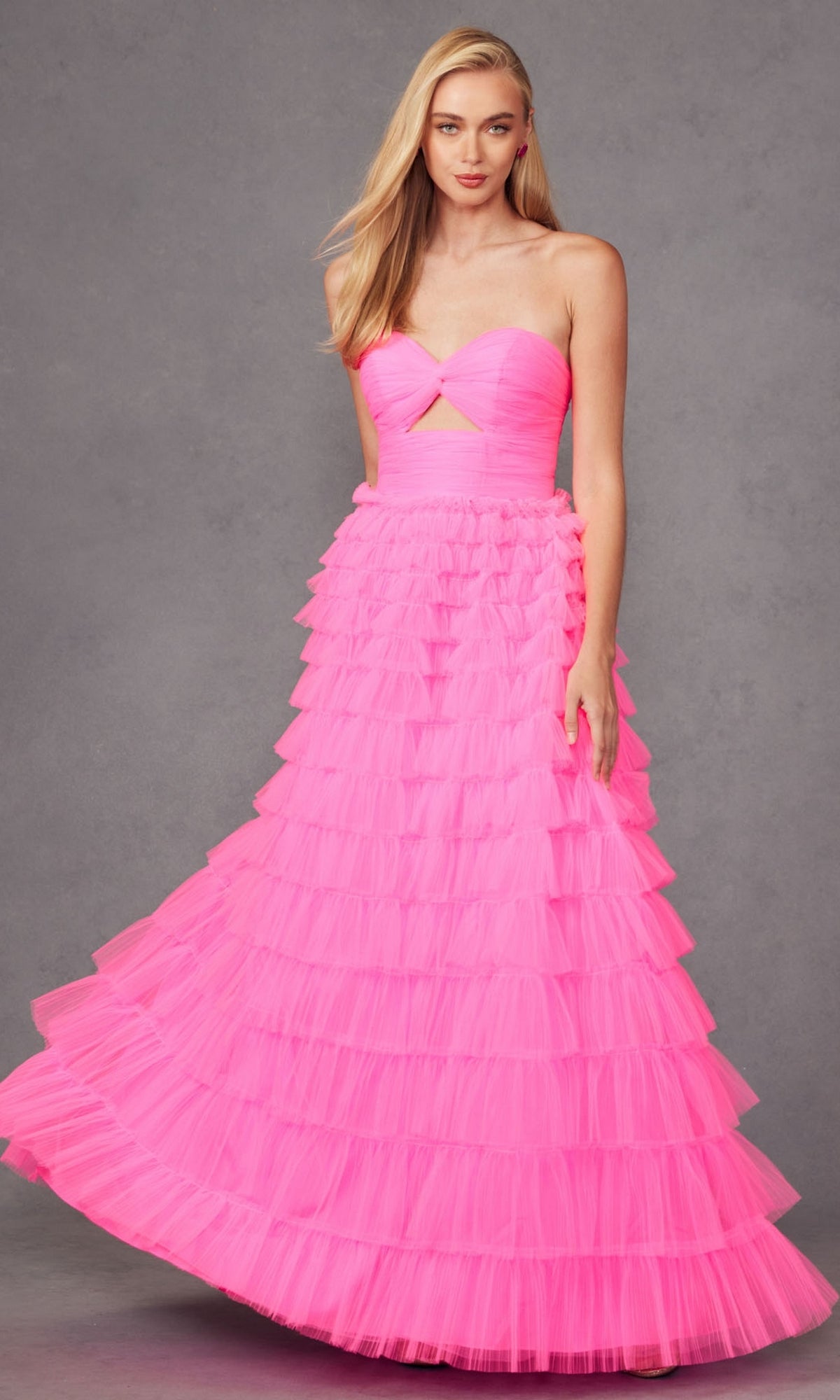 A woman wearing the Long Prom Dress JT2456H by Juliet—a strapless, bright pink ruffle tulle ball gown with a small front cutout and tiered layers—stands against a gray backdrop.