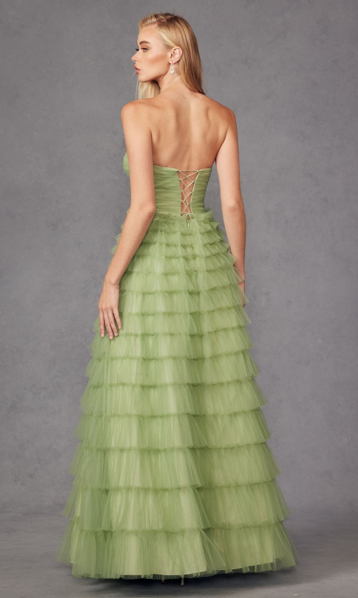 A woman faces away, wearing the Long Prom Dress JT2456H by Juliet—a strapless light green ruffle tulle ball gown with a lace-up back—set against a plain gray background.