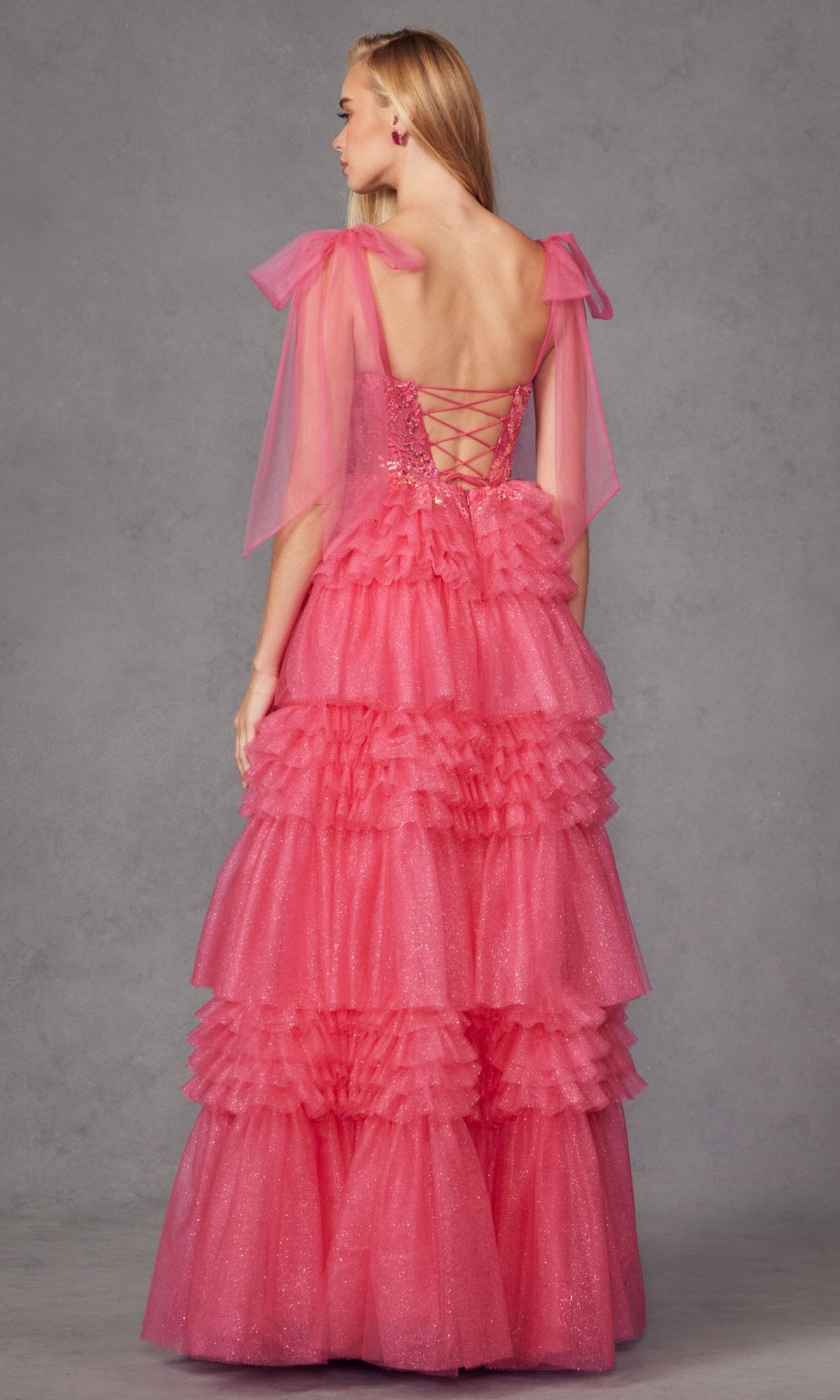 A woman in the Juliet JT2465A Long Ruffled Prom Ball Gown—featuring a glittery pink tiered skirt, sheer shoulder bows, and lace-up corset back—stands against a plain grey background.