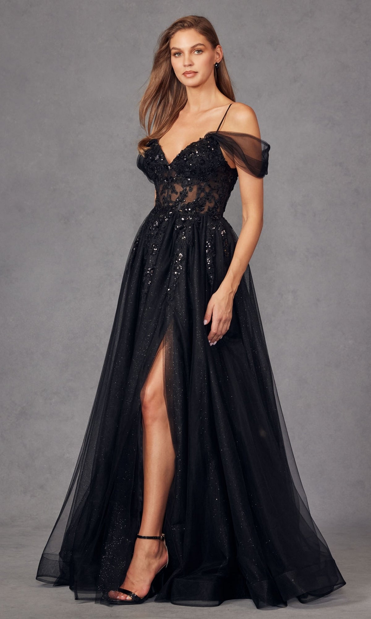 A woman models the Sheer-Waist Long Prom Ball Gown Juliet JT2466A—an elegant black, off-the-shoulder dress with a sheer bodice, floral accents, high slit, and spaghetti straps—against a plain gray background.