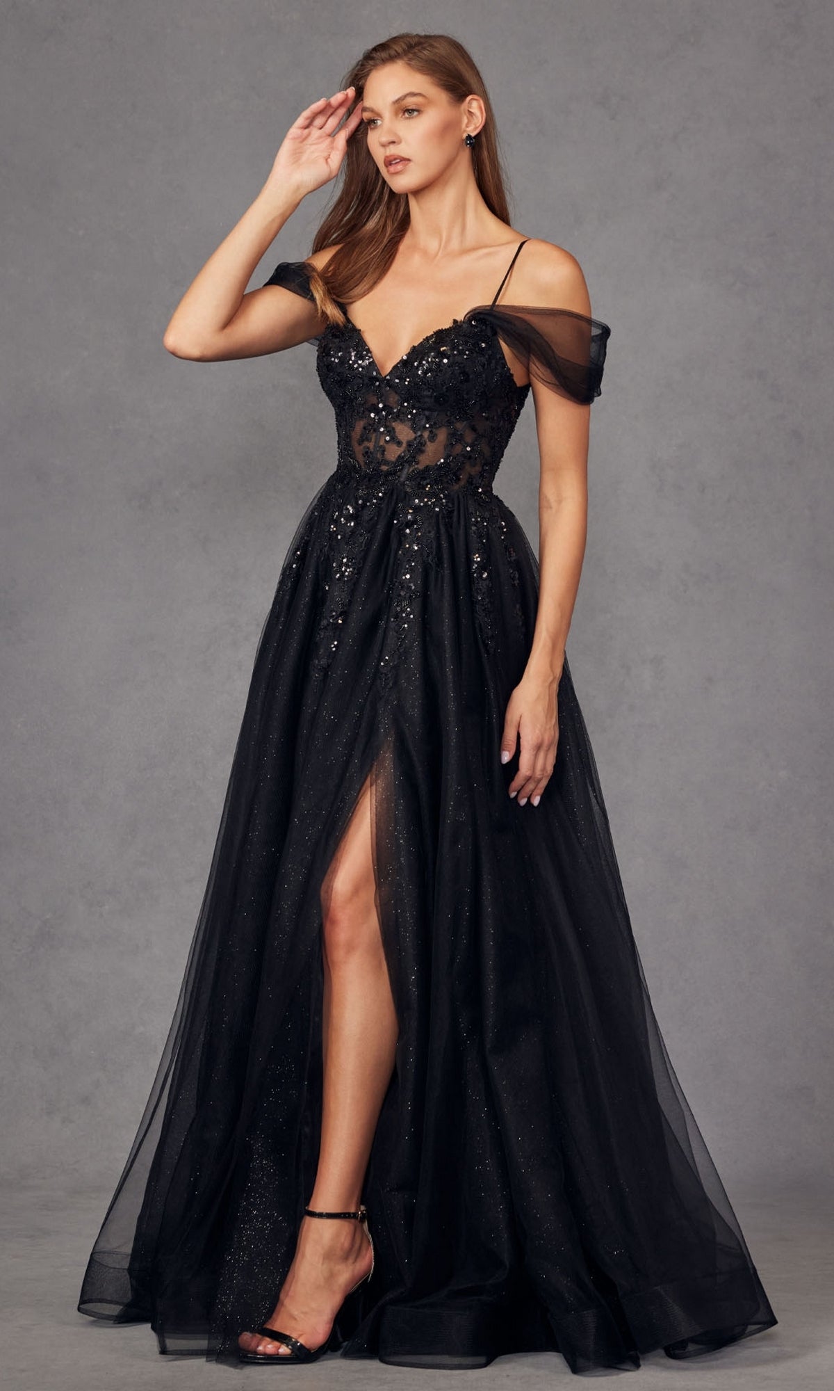 A woman in the Sheer-Waist Long Prom Ball Gown: Juliet JT2466A—black, off-the-shoulder with sequin and sheer details, thigh-high slit—pairs it with black high-heeled sandals, standing before a gray background.