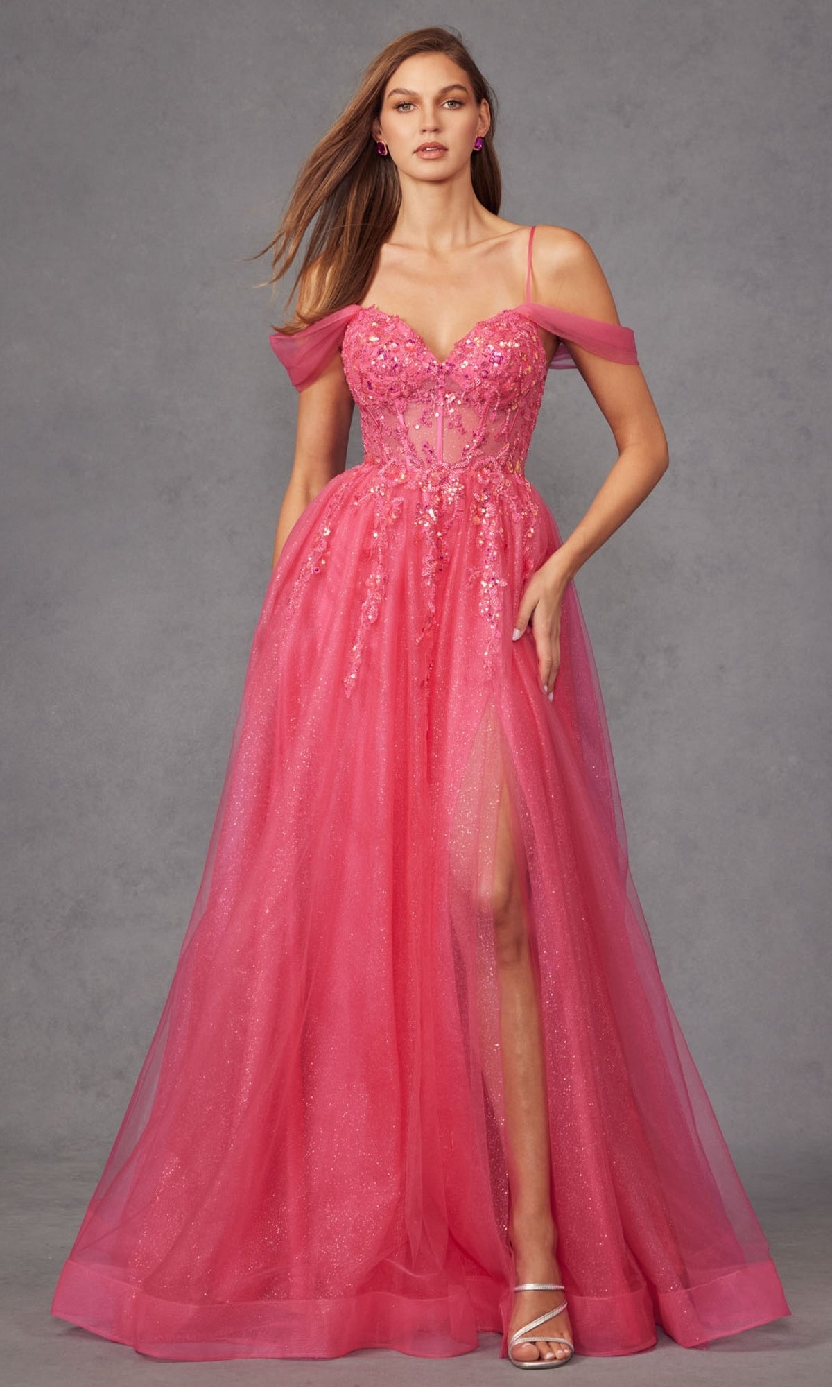 A woman in the Sheer-Waist Long Prom Ball Gown: Juliet JT2466A, a sleeveless pink floor-length dress with floral details, sheer off-shoulder straps, thigh-high slit, and strappy heels, stands against a gray background.