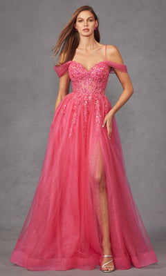 A woman in the Sheer-Waist Long Prom Ball Gown: Juliet JT2466A, a sleeveless pink floor-length dress with floral details, sheer off-shoulder straps, thigh-high slit, and strappy heels, stands against a gray background.